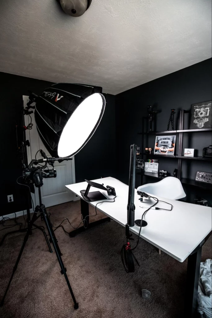 A small, dark-coloured office. There is camera gear scattered around the room, with a big studio light being used to light the area
